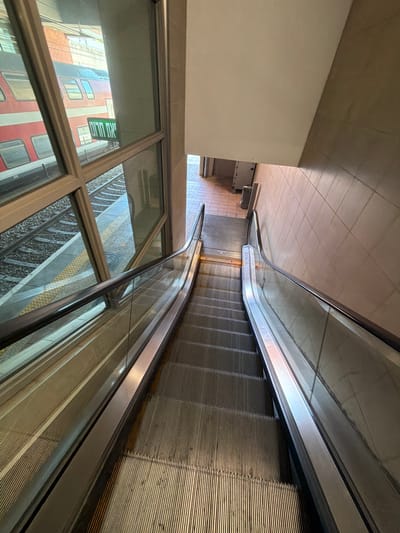 The image depicts an escalator leading down in a station, potentially underground. The escalator has metal treads and a glass side panel, through which a view of a railway platform is visible. The reflection in the glass shows a red and white building with multiple windows and a green and white sign, which indicates this may be the metro station in Holon, Israel. The station has tiled walls and flooring. The scene appears to be unoccupied.