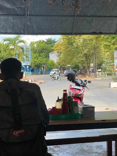 In Banjarbaru, Indonesia, a person wearing a backpack with an "OKKY" logo is seated at a table, looking out a window. The backpack is dark in color. In front of the person, on the table, is a green tray holding various items, including bottles of condiments (one red, one clear) and what appears to be a tissue box.
Through the window, a street scene is visible. A red motorcycle with a black helmet on its handlebars is parked in the foreground. The license plate reads "DA 6972 PBR". Further down the street, a green scooter with a rider wearing a white outfit is in motion. In the background, there are trees, buildings, and other vehicles. One building appears to be blue, with a white structure adjacent to it. A blue traffic sign with a white arrow pointing right is visible on the left side of the street. The sky is bright and appears to be daytime. The overall impression is that of a casual setting, possibly a roadside eatery or a rest stop.
