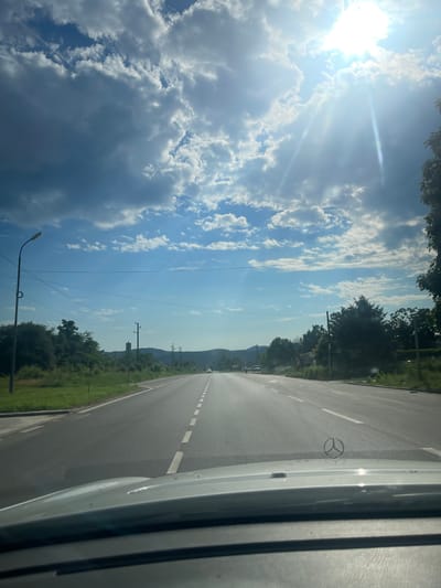 The image shows a view from the interior of a vehicle, looking out onto a straight, paved road in the city of Berkovitsa, Bulgaria. The road has white dashed lines marking the lanes. On the left side of the road, there is a grassy area and trees. On the right side, there are also trees and vegetation. The sky is blue with scattered clouds and the sun is shining brightly, casting rays of light. There are power lines and a lamppost visible along the road. In the distance, there are some buildings and hills. A Mercedes-Benz logo is visible on the hood of the vehicle in the foreground. There are no people clearly visible in the image.