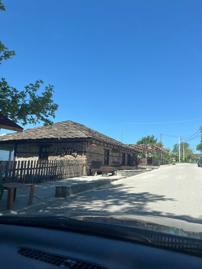 The image depicts a building in Leshten, Bulgaria, taken from inside a car. The architecture of the building is characterized by stone walls, a thatched roof, and a wooden fence. A sign with Cyrillic lettering is visible on the building. The sky is clear and blue, and tree branches with green leaves appear in the lower left corner of the frame. A road runs alongside the building, with shadows cast by trees. The interior of the car, including the dashboard and vents, is visible in the foreground.