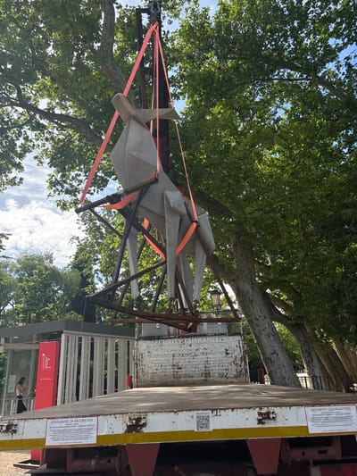 The image, originally captured in portrait orientation and currently rotated 90 degrees clockwise, presents an outdoor scene under bright, sunny daylight. The central subject is a large, abstract sculpture composed of angular, light grey concrete or stone forms. It is secured by multiple vivid orange cargo straps to a dark, open-framework metal structure, indicating that it is either in transit or being positioned for installation/deinstallation.
Above the sculpture, a dense canopy of lush green leaves from mature trees dominates the upper part of the frame, with glimpses of a clear blue sky and scattered white clouds visible through the foliage.
To the right, a portion of a flatbed truck or trailer is visible. Attached to its side are white signs with text and QR codes. Key inscriptions include "SECURITY GUARANTEES," "GARANZIE DI SICUREZZA," "Padiglione Ucraina," and "Pavilion of Ukraine at the 61° International Art Exhibition - La Biennale di Venezia." Another sign displays a "PRESS KIT" QR code. These details firmly place the setting at the Venice Biennale, specifically at the Ukraine Pavilion.
In the background, behind the truck, parts of a building entrance or gate can be seen, characterized by red accents. A person is discernible near this entrance, adding a human element to the scene. The overall impression is one of active preparation or handling of a significant art installation within the context of a major international exhibition in Venice, Italy. The weather appears pleasant and clear.