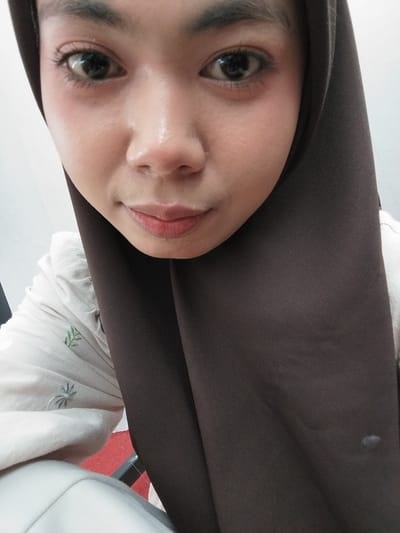 The image is a close-up, frontal portrait of a young woman wearing a dark brown hijab and a light-colored shirt with subtle green embroidery. She is looking directly at the camera with a neutral expression. The focus is on her face, with her eyes, nose, and lips clearly visible. The background appears to be an indoor setting with a white wall. The lighting is even, highlighting the contours of her face and the texture of her hijab. The image is shot from a slightly high angle, looking down at the subject. There is no visible indication of the location being Tibojong, Indonesia.