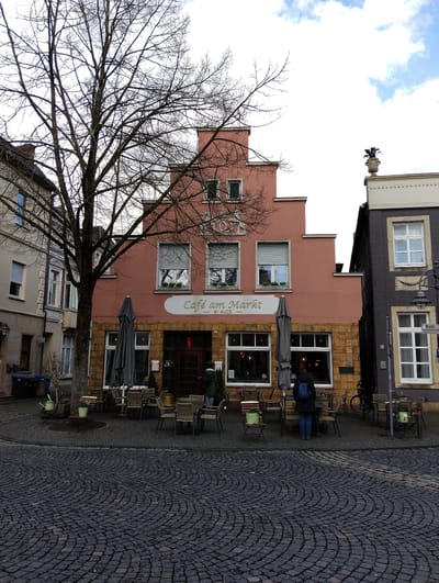 The image depicts a street scene in Burgsteinfurt, Germany, specifically around a market square. The central focus is "Café am Markt," a two-story building featuring a distinctive pinkish-red facade with a stepped gable roof and an ochre-yellow stone block ground floor. The cafe's entrance consists of brown double doors with glass panels. Above the entrance, a white oval sign reads "Café am Markt," with "BY ALICE" in smaller text below it.
Outdoor seating arrangements, including numerous small square tables and matching chairs made of a wicker-like material, are present on the dark grey cobblestone pavement, which is laid in a fan pattern. Two large grey folded umbrellas are positioned over the seating areas. Several small green potted plants are used as decoration on tables and around the base of a prominent, leafless deciduous tree that stands centrally in front of the cafe.
To the left of the cafe, a lighter beige/cream building with multiple windows and roller shutters is visible. To the right, a dark grey building with white-framed windows features a decorative urn-like planter on its roofline. A lamppost with a "Markt" street sign is situated near this building.
One individual, wearing a dark jacket and a blue backpack, stands with their back to the viewer, facing the cafe's entrance. Another person is partially visible near the cafe's doorway, seemingly engaging in an interaction. A black bicycle is parked near the cafe's right side, and two black refuse bins are visible to the far left. The sky overhead is partly cloudy, displaying patches of blue and white clouds.