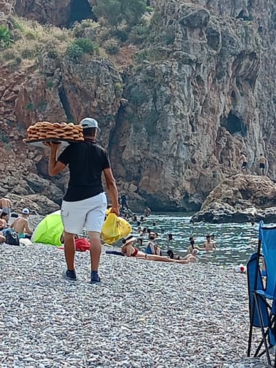 A man walks along a rocky beach in Antalya, Turkey, carrying a tray of baked goods on his shoulder and a yellow bag in his hand. The beach is populated with many people swimming in the clear blue water and sunbathing on the shore. Towering, rugged cliffs with sparse vegetation form a dramatic backdrop to the scene. The water is calm, with small ripples indicating gentle movement. Some beach chairs are visible in the foreground, suggesting a tourist-oriented location. The man, dressed in a dark polo shirt, white shorts, and a cap, appears to be a vendor offering his products to beachgoers.