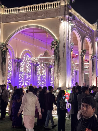 A wedding reception at night shows guests standing before a multi-tiered architectural structure adorned with flowers and delicate lighting. Arches define each level, revealing ornate latticework and sheer drapery. The crowd, dressed in formal attire, is an active audience. A uniformed individual is visible near the top of the frame. The event has a lavish and celebratory ambiance.