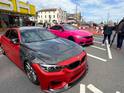The image shows a street scene in Great Yarmouth, United Kingdom. Two customized BMW cars are parked on the street, which is also being used as a pedestrian thoroughfare. The car in the foreground is a red-and-black custom model, featuring a black hood and carbon fiber detailing on the side panels. Behind it, a pink BMW stands out against the backdrop. Pedestrians, some partially visible, are walking along the street, separated from the cars by temporary orange barriers. Shops with yellow awnings and signs reading "TOYS - CASH PRIZES" line the street on the left. The architecture in the background includes buildings with white facades and multiple windows. Union Jack flags adorn the streetlights. The sky is partly cloudy.