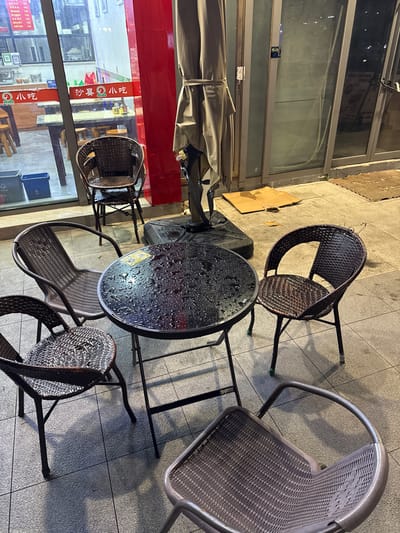 This image depicts an outdoor seating area in front of a business located on Nanjing Road Pedestrian Street in the Huangpu District of Shanghai, China. A small, round, dark-colored table with visible water droplets sits in the center of the frame, suggesting recent rain. Four woven chairs surround the table. Two chairs are dark brown rattan, and two appear to be a lighter brown rattan. A fifth, metallic gray chair is partially visible in the lower right corner of the image. Behind the table and chairs, a large, light brown umbrella is folded and tied up. Through the glass windows of the establishment, tables and chairs are visible inside, along with signage in Chinese. Two blue recycling bins are positioned near the window to the left. The ground is paved with light gray tiles, and shadows are cast by the table and chairs. The overall scene suggests a temporarily closed outdoor dining or waiting area.