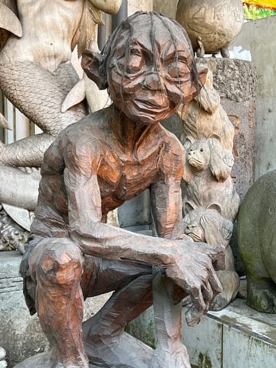 The image shows several wooden carvings displayed outdoors, likely for sale, in Ubud, Indonesia. The carvings include a dark brown, roughly-hewn human-like figure with its arms raised; a lighter colored carving of several monkeys huddled together; a dragon-like figure with scales and fins; and other various carved stone or wood objects in the background. The carvings are sitting on or leaning against a stone or concrete surface. The overall setting appears to be a shop or market area.