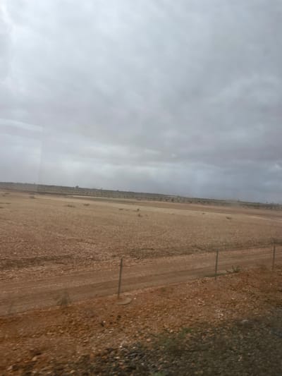 A flat, arid landscape stretches under a cloudy sky. The ground is predominantly brown and rocky, suggesting a desert or semi-desert environment. A rough dirt road cuts through the terrain, marked by tire tracks. Thin poles, possibly fence posts or utility poles, line the road. Vegetation is sparse and scattered. The overall scene conveys a sense of desolation and remoteness.