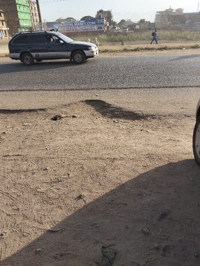 This image displays a roadside scene in Thika, Kenya, under bright daylight. A dark blue and silver Toyota G-Touring station wagon, equipped with a roof rack, moves rightward on a paved asphalt road. The driver is visible within the vehicle. To the right of the moving vehicle, a single individual in blue clothing walks eastward on a dirt verge parallel to the road. In the background, a large billboard for "ROYAL MABATI.COM" advertises roofing sheets with contact numbers 0722 63 63 63 and 0720 04 04 04. To the far left, a multi-story building under construction, partially covered in green netting, is visible. Other urban structures and sparse vegetation are present in the middle and far background. The foreground consists of a dry, light-brown dirt ground displaying tire tracks, footprints, scattered small stones, and unidentifiable dark debris. Shadows extend across the dirt, indicating a low sun angle. A section of a vehicle's front tire and fender is visible on the extreme right edge of the frame, from which the photograph appears to have been taken.