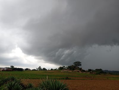 The image depicts a rural landscape under a dramatic, heavily clouded sky, suggestive of an impending storm. The foreground features cultivated fields with brown, tilled earth and rows of green plants, possibly crops. In the midground, there are more green fields, some appearing to be newly planted, and a cluster of structures that resemble small buildings with tin roofs. Further back, a rocky outcrop is visible, topped with a large, mature tree and other vegetation. Two figures, likely farmers, are present in the fields, one walking and the other tending to the land. The background consists of rolling hills and a dense, dark cloud formation that dominates the sky, obscuring the sun and casting a shadow over the entire scene. The location is described as Katon Rikkos, Nigeria.