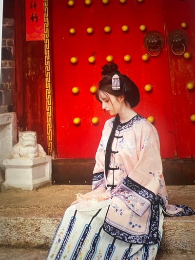 A person is posing in front of a red, ornate door. The door has several round, golden decorative elements and two darker, circular metal door knockers. The person is wearing a traditional Chinese garment with a pink base and blue and white floral patterns. The garment has a high collar and is fastened at the front. The person's hair is styled up and adorned with a decorative hair clip. The background consists of a textured, stone-like surface on the right and a wooden panel on the left, partially obscured by the person. Below the red door, there is a decorative border pattern and a red rectangular sign with yellow Chinese characters. The overall scene appears to be set outdoors, possibly in a historical or cultural site in Liandu District, China. The lighting suggests it could be daytime.
