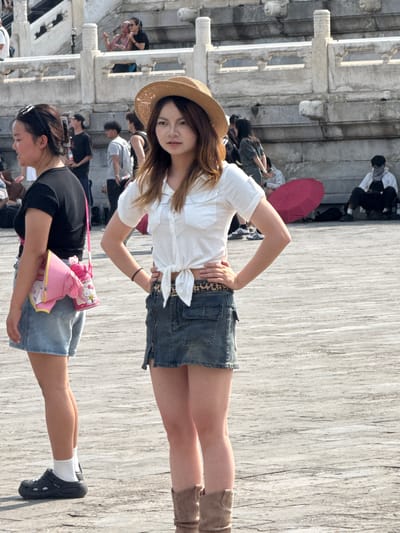 In Dongcheng District, China, two young women are standing on a paved outdoor area, possibly a plaza or courtyard, with a marble structure in the background. The woman on the left, wearing a black t-shirt, denim shorts, white socks, and black clogs, has a pink Hello Kitty-themed bag slung over her shoulder. The woman on the right, facing the camera, is wearing a white tied-up short-sleeved shirt, a denim mini-skirt with a leopard print belt, and tan boots. She has a straw hat on her head and is posing with her hands on her hips. Several other people are visible in the background, some sitting, some standing, and some walking, near the marble structure. A red umbrella is lying on the ground behind the woman on the right. The overall scene suggests a public gathering or tourist area.