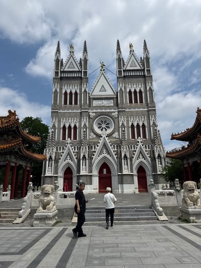 This is a photograph taken outside the North Cathedral, also known as the Church of the Immaculate Conception, located in Xicheng District, China. The cathedral is a grand Gothic-style building with twin spires, ornate stone carvings, and red double doors. In the foreground, two individuals are walking on a paved plaza. To the left and right of the cathedral entrance are traditional Chinese-style pavilions with orange tiled roofs and red pillars. Stone lion statues are positioned on either side of the entrance steps. The sky above is partly cloudy.