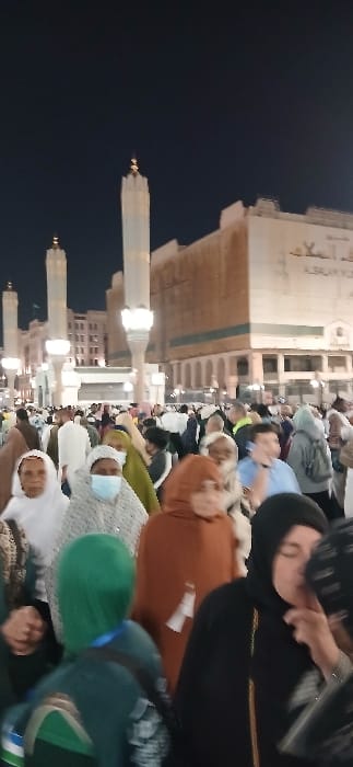 The image depicts a densely crowded outdoor plaza at night, located in the city of Medina, Saudi Arabia, adjacent to the Prophet's Mosque (Al-Masjid an-Nabawi). Multiple tall, slender, light-colored minarets with golden finials are prominently featured against a dark sky, illuminated by artificial light from below. In the background, multi-story buildings, including one with a light brown facade displaying Arabic script (partially visible as "السلام" or "Al-Salam"), are also lit.

The foreground and midground are filled with a large congregation of people, predominantly adults, moving or standing in close proximity. Individuals are dressed in various modest attires; many women wear head coverings such as white, green, black, and brown hijabs or abayas. One woman in the immediate foreground, wearing a patterned head covering, has a light blue surgical mask over her mouth and nose. Another individual in the foreground wears a green head covering and a backpack, facing away from the viewer. A woman in a black abaya and hijab is visible on the right foreground, with her left hand near her face. General actions include walking and standing within the crowd. The scene is illuminated by ambient artificial light sources, casting light on the structures and crowd.