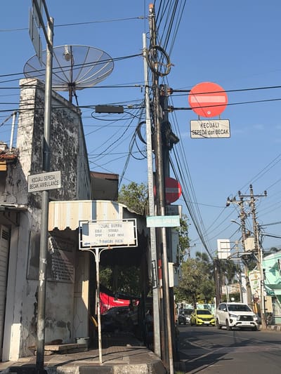 The image depicts a street scene in Pekalongan, Indonesia, under a clear blue sky. In the foreground, a weathered, light-colored building with peeling paint stands on the left, adjacent to a sidewalk. A metal pole with various signs is mounted in front of the building. One sign, written in white text on a black background, reads "KECUALI SEPEDA / BECAK/AMBULANCE." Further down the street, another pole displays a circular red sign with a white horizontal bar, indicating "No Entry," accompanied by a smaller rectangular sign below it stating "KECUALI / SEPEDA dan BECAK" (Except bicycles and pedicabs).

To the right of the street pole is a building with a striped awning. A white sign hangs from a pole in front of this building, listing "JUAL BUMBU" (Spice Seller) and various spice names. Behind this, a red tarp is visible.

The street itself is paved and has lane markings. Several vehicles are present: a white SUV, a yellow hatchback, and a dark-colored car further back, all moving in the same direction. Utility poles with numerous wires are scattered throughout the scene, indicating a busy urban environment. In the background, more buildings and trees are visible. The overall impression is a typical street view in a densely populated area of Indonesia.