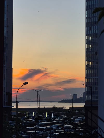 The image depicts a panoramic view of the Amur River at sunset, as seen from the city of Khabarovsk, Russia. In the foreground, two modern high-rise buildings frame the scene, one on the left and one on the right. Between them, a large parking lot is filled with numerous cars. The river occupies the middle ground, with a promenade or embankment visible along its edge where several figures are gathered. Distant city buildings are silhouetted against the colorful sunset sky, which transitions from a pale yellow at the top to vibrant oranges and pinks lower down. A lone boat is visible on the water. Streetlights are present along the embankment and within the parking lot. The overall atmosphere is one of urban tranquility at the close of day.