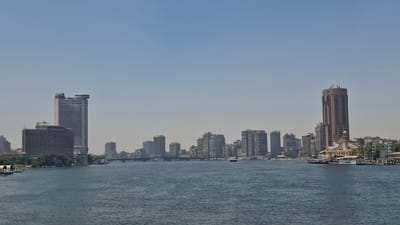 This is a wide-angle photograph of a cityscape viewed across a body of water. The scene depicts a collection of modern skyscrapers and other buildings lining the distant shore. The water in the foreground is a deep blue-gray, with gentle ripples suggesting a calm day. Several boats of various sizes are visible on the water, including a few larger passenger vessels docked or moving slowly near the shore.
The sky is a pale, hazy blue, indicating a clear but potentially warm day. The light suggests it is daytime, likely late morning or early afternoon, given the brightness and the lack of strong shadows.
On the left side of the image, a tall, light-colored building with a distinctive rounded top is prominent, bearing the word "Hilton" near its peak. Next to it is a darker, more block-like building. Further along the skyline are numerous other high-rise structures of varying heights and architectural styles, creating a dense urban backdrop.
On the right side of the image, a tall, reddish-brown cylindrical building stands out. The word "SOFITEL" is visible near the top of this structure. Below it, a circular, cream-colored building with a domed roof and ornate detailing is situated on the waterfront, with several boats docked alongside it.
The overall impression is of a bustling, modern city situated along a significant waterway, likely a river. The distant buildings and the presence of the water suggest an urban environment with a connection to commerce and transport. There are no visible people in the frame. The atmosphere appears serene and somewhat distant, characteristic of a panoramic cityscape.