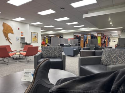 A library lounge area features various seating options, including orange chairs around a small table, and gray armchairs with patterned pillows. Bookshelves line the background. A wall display includes an abstract wooden sculpture, a water fountain, and a framed poster. In the foreground, a person's leg wearing a black boot rests on a table, next to a promotional pamphlet. The room is brightly lit by recessed ceiling lights.