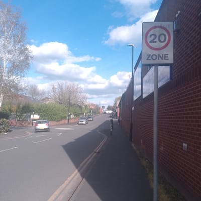 The image depicts a street scene in what appears to be a residential or mixed-use area. A "20 ZONE" speed limit sign is prominently displayed on the right side of the frame, affixed to a brick building. The street slopes upwards and away from the camera, with parked and moving vehicles visible in the distance. A person is walking on the sidewalk on the right side of the street. Trees and shrubbery line the left side of the road. The sky is blue with scattered white clouds. This image, with the "20 ZONE" speed limit sign, is located in Coventry, United Kingdom.