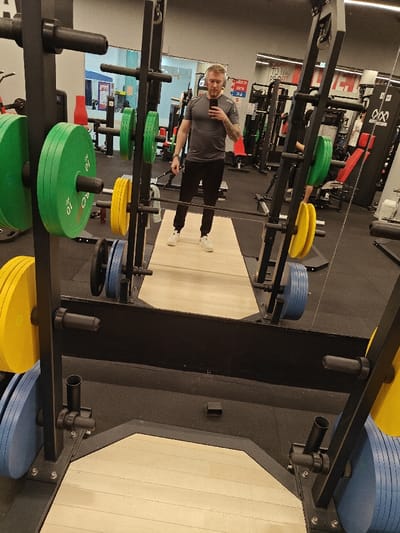 A mirrored view within a gym setting showcases a man standing on a wooden platform, holding a cell phone. He is dressed in a grey t-shirt and dark pants, and he appears to be taking a selfie. The squat rack in the foreground is loaded with various colorful weight plates. Other gym equipment is visible in the background, alongside gym branding.