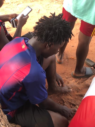 The image depicts a casual outdoor gathering of at least three individuals on reddish-brown soil, typical of the natural environment in Nsugbe, Nigeria. The scene is brightly illuminated by natural sunlight, indicating it is daytime.

In the foreground, a person with dark dreadlocks is seen from behind, wearing a distinctive blue and red striped shirt. This individual is actively engaged with a white smartphone, holding it with both hands and appearing to interact with the screen. A cable extends from the smartphone, possibly connecting to a charging device or power bank.

Other people are partially visible around the central figure. To the upper left, one person wears a light green top and red shorts adorned with yellow stripes. Another individual's bare feet and lower legs are visible on the upper right, wearing black shorts, with a dark grey flip-flop resting on the ground nearby. A glimpse of red and white fabric on the far right suggests the presence of a fourth person.

The ground consists of coarse, reddish-brown dirt or sand, with scattered small organic debris such as twigs and leaves. The overall atmosphere appears relaxed and informal, suggesting a social gathering in a warm climate. The casual clothing and bare feet contribute to this impression. While the individuals' emotions are not discernible, the activity centers around informal interaction and the use of a smartphone. No specific readable text is visible on the phone or clothing, beyond decorative yellow stripes on one shirt.