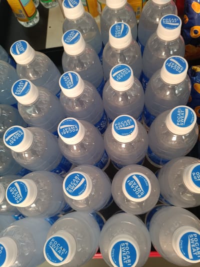 The image shows a close-up, overhead view of numerous bottles of Pocari Sweat, a Japanese sports drink. The bottles are arranged in multiple rows, filling the frame. Each bottle has a white cap with a blue and white Pocari Sweat logo, and the word "OPEN" is visible on many of the caps. The bottles themselves are clear plastic and appear to contain a light, clear liquid, some with condensation on the outside, suggesting they are cold. The lighting is somewhat dim, and the background is a dark surface, possibly a shelf or table, with a hint of other products to the left and right. The overall impression is of a retail display or stock of beverages. There is no visible human presence. The setting appears to be indoors, likely a store or warehouse. The image is still and does not capture any specific activity or event beyond the static arrangement of the products. The visible text is primarily the brand name "Pocari Sweat" and the instruction "OPEN" on the bottle caps. The location context provided is Depok, Indonesia.