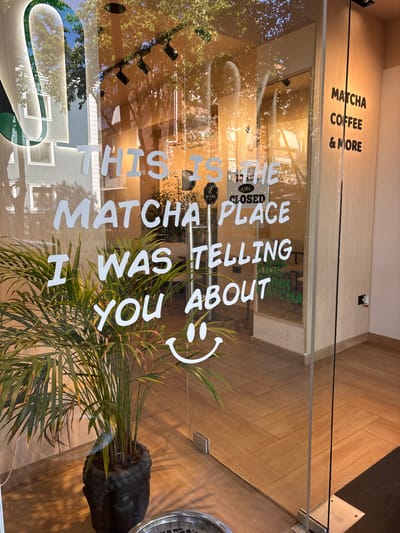 Here's a description of the image:
The image captures the entrance of "100% Organic Matcca" in Tirana, Albania. A glass door displays the words "THIS IS THE MATCHA PLACE I WAS TELLING YOU ABOUT" with a smiley face, and a "PULL TO OPEN" sign. The door also has a "CLOSED" sign visible.
Above the door, a sign reads "MATCHA COFFEE & MORE". Reflections on the glass door reveal a street scene with trees and buildings. A potted plant is positioned near the entrance on the interior. A Buddha head pot holds the greenery. Tiled wood-look flooring is visible.