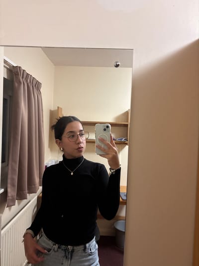 A person in a black turtleneck and light wash jeans takes a mirror selfie. They hold an iPhone in their right hand, displaying the back of the phone. The person wears round glasses and a delicate necklace. Behind them, drawn tan curtains hang over a window. A beige wall and minimalist shelving are visible in the reflection.