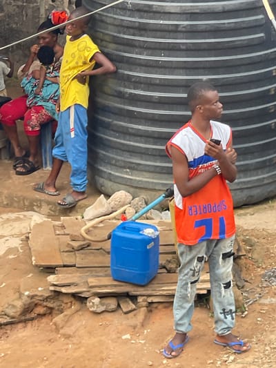 Two people stand near a large corrugated metal tank, one wearing a colorful shirt with "SUPPORTER" on the back and ripped jeans, the other in a yellow shirt and blue pants. A blue jerrycan sits on wooden planks with a hose leading to the tank. Below, a woman in a floral print shirt and red pants crouches, and a child in yellow and blue stands beside her. Another person is partially visible, wearing a white shirt. The ground around them appears to be a mix of dirt, wood, and rocks.