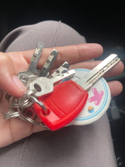 A person's hand, exhibiting faint fingerprint patterns and a light skin tone, is holding a set of keys. The keys are attached to a circular keychain that features a white background with blue trim, and a pink letter "K" with a blue and orange smiling face and a smaller orange object on it. One of the keys has a red plastic head. The background consists of dark fabric, likely clothing, which appears to be greyish-brown. The image is taken from a close-up perspective, focusing on the hand and keys. There are no visible indicators suggesting this scene is located in South Tangerang, Indonesia.