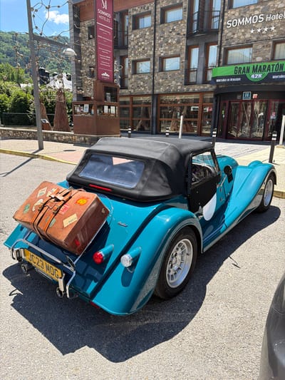 In the city of Benasque, Spain, a teal-colored classic convertible sports car is parked on an asphalt surface outside a stone-faced building. The car has a black convertible top. A vintage brown suitcase, adorned with multiple travel stickers, is secured to the rear of the car. The car's license plate reads "JC23 MOG". The building in the background displays signage for "SOMMOS Hoteles" and "GRAN MARATÓN 42K Montañas de Benasque". The scene is illuminated by daylight, casting shadows on the ground.
