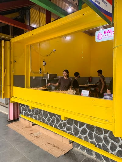 The image shows a food stall in Malang, Indonesia, located at coordinates -7.968, 112.628. The stall has a bright yellow exterior with a stone base. A woman is inside the stall, preparing food with a spatula. There are two other people in the background, one standing and another sitting. The stall has a sign that says "HALAL INDONESIA" with a phone number. The scene is likely a street food stall, offering various food items. The image is taken from a slightly elevated angle, showing the stall's exterior and the surrounding area.