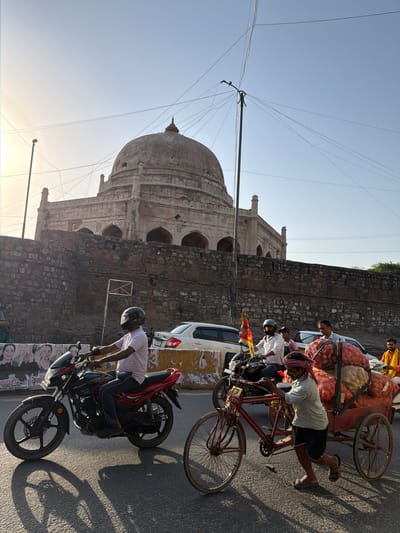 This image depicts a street scene in Delhi, India, with a prominent dome and stone wall of an old building on the left. Several people are engaged in various activities on the right side of the frame. In the foreground, a man is sitting on a red motorcycle, facing away from the camera. Behind him, a man is leaning over to push a cart laden with large sacks of produce, possibly onions, which is attached to a bicycle. Another man is standing next to the cart, looking towards the street. Further back, more people are visible near other vehicles, including a white car and another cart. The scene appears to be taking place during the day, with the sun casting long shadows on the asphalt road. The sky is clear and blue, suggesting fair weather. The presence of the ancient architecture alongside the bustling street activity provides a glimpse into the blend of historical and modern life in Delhi. There is no visible text in the image.