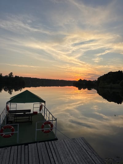 The image depicts a serene sunset over a calm body of water, likely a lake or river, near the city of Kamysh, Russia. In the foreground, a wooden pier leads to a small, covered dock. The dock is equipped with two benches and a table, suggesting it is a resting or gathering spot. Two orange life preservers are attached to the railing of the dock, indicating a potential for water activities or safety measures. The water reflects the soft hues of the sunset, creating a mirrored image of the sky and the surrounding treeline. The sky itself is filled with wispy clouds painted in shades of orange, pink, and pale blue, with the sun setting on the horizon. The treeline forms a dark silhouette against the vibrant sky, suggesting a natural, possibly forested, landscape surrounding the water. No people are visible in the image. The overall atmosphere is tranquil and picturesque, capturing the beauty of a natural sunset in a rural or semi-rural setting.