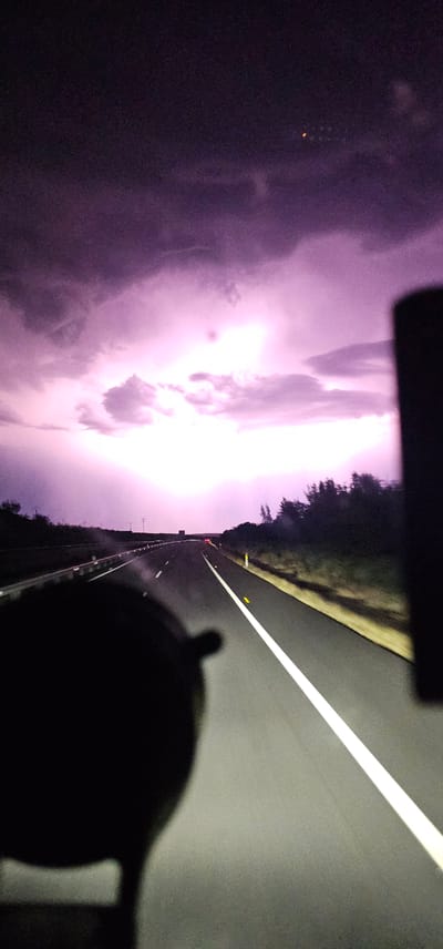 The image depicts a view from inside a vehicle traveling on a highway at night. The sky is dominated by a dramatic display of lightning, casting a bright pink and purple glow on the clouds and the surrounding landscape. The highway stretches forward, with a white line marking the center and yellow markers on the right side. On the left side of the road, a guardrail is visible, and beyond that, a dark, hilly terrain with sparse vegetation. On the right side, a dense line of trees forms the edge of the highway. In the foreground, a dark, out-of-focus object, likely part of the vehicle's interior or an accessory, obscures the lower left portion of the frame. The lighting suggests an active thunderstorm. The location is identified as being in the vicinity of Malpartida de Plasencia, Spain.