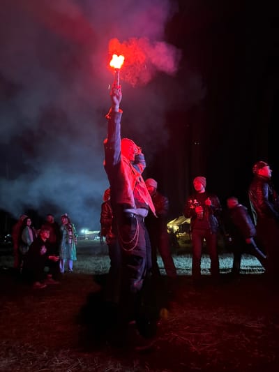 The image displays an outdoor nighttime gathering in Большая Кивара, Russia. A central figure in the foreground, wearing dark clothing and a chain accessory, stands with their right arm extended vertically, holding a bright red handheld flare that emits dense red smoke. The flare's intense light casts a strong red illumination over the immediate surroundings and the individuals present.
Approximately 10-15 people are observable in the midground and background. To the left of the central figure, a person in a dark hoodie is crouching, while another person stands nearby, clad in a translucent green poncho. Further left, a male figure with a beard is visible, illuminated by the red light, amongst other less distinct individuals. To the right of the central figure, a man wearing a dark jacket and a light-colored beanie stands holding a clear plastic bottle. To his right, another man in a dark leather jacket and a dark cap is positioned, looking towards the right side of the frame. A blurred figure is also visible on the far right, suggesting motion.
The ground consists of an unpaved area covered in sparse grass or straw, uniformly colored red by the flare's light. In the distant midground, a white tent-like structure is discernible, emanating a soft yellow internal light. The background features dark silhouettes of trees against the night sky. Red smoke from the flare is distributed throughout the scene, creating a hazy atmosphere, particularly noticeable around the flare and in the upper left quadrant of the image.