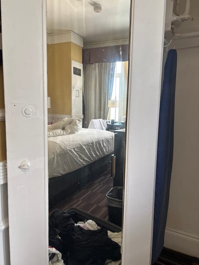 The image shows a full-length mirror reflecting a messy hotel room. The reflection displays a bed with rumpled white linens, a nightstand with a lamp and bottles, and a window with sheer curtains. A suitcase overflowing with clothes is visible in the foreground of the reflection, along with a trash can. To the right of the mirror, a dark blue garment hangs on a hook, and a white ironing board is visible above it. The room appears to be in a state of disarray, suggesting recent use or packing. The overall impression is of a lived-in and somewhat untidy hotel room.