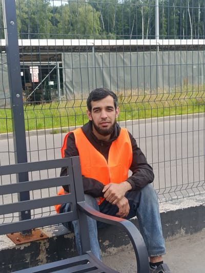 A man wearing an orange safety vest and blue jeans sits on a bench in front of a chain-link fence. Behind the fence, trees and a building are visible. The scene appears to be outdoors, possibly in a construction area or industrial site in the city of Kommunarka, Russia. The man is looking directly at the camera with a neutral expression.