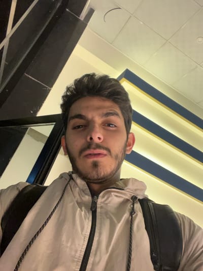 A young man with dark hair and a beard poses for a selfie indoors. He is wearing a light-colored jacket and a backpack. The background features a ceiling with recessed lighting and blue and gold accents. A black metal railing is visible on the left side of the frame. The photograph is taken at a slightly tilted angle, looking upwards at the subject.