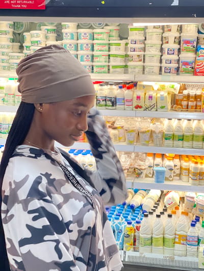 The image shows a young Black woman in a grocery store, likely in Guzape, Nigeria, based on the provided context. She is wearing a headscarf and a camouflage-patterned jacket with the words "WORSHIPHEROWOR" visible on a white strap across the shoulder. The woman is looking to her right, with a neutral expression.
Behind her, the scene is a brightly lit refrigerator aisle filled with various dairy products. Numerous containers of yogurt, labeled "GREEK" and "HABIB," are stacked neatly. There are also several bottles of milk, some with blue caps and labels, and other assorted beverage containers. Some of the bottles appear to be from brands like "Amira" and "Amul." The overall impression is one of a well-stocked grocery store refrigeration section. A red sign with white text that reads "ARE NOT RETURNABLE THANK YOU" is visible on the left side of the image. The lighting suggests it is daytime inside the store.