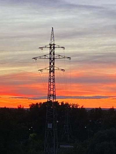 A silhouette of a high-voltage electricity pylon stands against a vibrant sunset sky. The sky transitions from a pale gray at the top to fiery oranges and reds near the horizon, with distinct cloud formations visible. The pylon, made of dark metal latticework, is topped with insulators and wires that stretch out of frame. Below the pylon, a dense line of dark green trees forms the horizon, obscuring the ground. The overall scene depicts a somewhat industrial landscape meeting natural beauty at dusk. The specific location in Novokuybyshevsk, Russia, is an area with power infrastructure amidst natural surroundings.