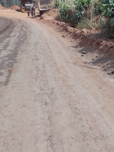 The image depicts an unpaved, reddish-brown dirt road in Hoche, Nigeria, under daylight conditions. The road surface shows scattered small stones and faint vehicle tracks, curving slightly to the left in the distance. To the right of the road, an elevated embankment of excavated reddish-brown earth supports green grasses and shrubs. A thin, light-colored linear object lies on the road in the mid-ground. In the upper-left background, two large heavy vehicles are visible: a yellow dump truck or construction machine, and a dark-colored dump truck positioned immediately to its right. Two individuals are present near the yellow vehicle; one is pushing a small two-wheeled cart, and the second stands adjacent. Distant green foliage constitutes the far background.