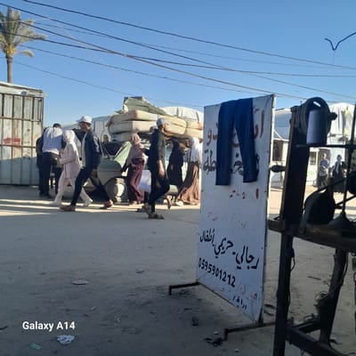 The image, taken in the city of Al-Mawasi, Unknown, shows a street scene. A group of people are walking on the street in the vicinity of what seems to be a parked truck or a vehicle loaded with mattresses and other items. The truck is partially visible in the lower part of the frame. Along the street, there's a sign, appearing to be advertising clothing, with some Arabic writing and a phone number (0595901212) visible. Power lines are visible in the sky.