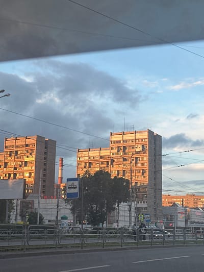 The media file shows a street scene in Magnitogorsk, Russia, likely during late afternoon or early evening. The perspective is from inside a vehicle, looking out at a series of tall, Soviet-era apartment buildings. The buildings are concrete structures with numerous windows, many of which are illuminated by the setting sun, casting a warm glow.
In the foreground, a grey, curved surface, possibly a vehicle window or a building facade, occupies the left side of the frame, bisected by a diagonal line of light and shadow. Overhead power lines crisscross the sky, a common feature in urban Russian landscapes.
The street itself is visible on the right, with a grey asphalt road and white lane markings. A metal fence separates the sidewalk from the road. On the sidewalk, there are several people, some standing and others seated, appearing to be waiting. A street sign with a pictogram of a bus and a yellow diamond-shaped sign with a black symbol are also visible.
Behind the fence, further buildings and trees are visible. One prominent feature is a tall industrial chimney with red and white banding, likely a landmark of Magnitogorsk, a city known for its heavy industry. Signs with Russian text are present on some of the buildings, though they are partially obscured and difficult to read clearly. The sky is a mix of blue and grey clouds, indicating overcast conditions with patches of sunlight. The overall mood is that of a typical urban evening, with the setting sun adding a touch of warmth to the otherwise utilitarian architecture.