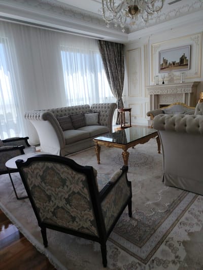 A formal, well-decorated living room features ornate white molding on the ceiling and walls, a large crystal chandelier, sheer white curtains, and patterned drapes. A plush, light-colored tufted sofa with decorative pillows sits on a large, patterned rug. A dark wood and gold-trimmed glass coffee table is in front of the sofa. To the left of the sofa is a dark wood armchair with a patterned upholstery. A fireplace with decorative stonework and a framed artwork of a city skyline is on the right side of the room. This living room is located in a hotel in Konigil, Uzbekistan.