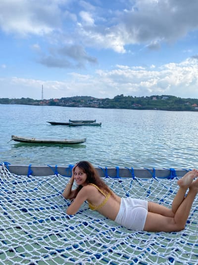 A woman lies on a white net hammock suspended over the ocean in Nusa Penida, Indonesia. She wears a yellow bikini top and white shorts. The ocean is calm with two small boats visible in the background. The shoreline, covered in lush green foliage and dotted with buildings, forms a gentle curve against a sky filled with scattered clouds. A telecommunications tower is prominent on the distant hills.