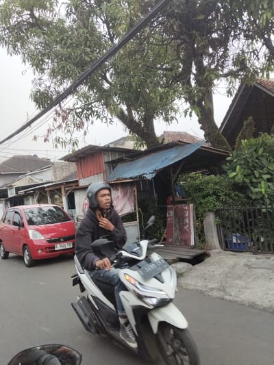 This media file captures a street scene in Bandung City, Indonesia. The primary subjects are a person riding a white scooter and a red car in the background. The scooter rider, wearing a dark hoodie and a helmet, is in the foreground, looking towards the left of the frame. The red car, a small hatchback, is positioned behind the scooter.
The setting appears to be a moderately busy street with buildings and trees lining the road. Overhead, power lines are visible, crisscrossing the frame. Some structures appear to be small shops or dwellings with corrugated metal roofs and wooden supports. Dense green foliage from trees and bushes is also prominent, suggesting a natural element integrated into the urban environment. The ground is paved with asphalt and some concrete.
The time of day is difficult to determine precisely, but the diffused lighting suggests it could be overcast or early morning/late afternoon. The weather appears to be neutral, without distinct signs of rain or strong sun.
A notable detail is the license plate on the red car, which reads "D 1624 YTP". Another license plate is partially visible on the scooter. A red sign with white text, partially obscured by a bush, is visible on the right side of the frame, with "NEW" and "AZER" being legible parts of the text. The overall atmosphere is that of an everyday urban scene in Indonesia.