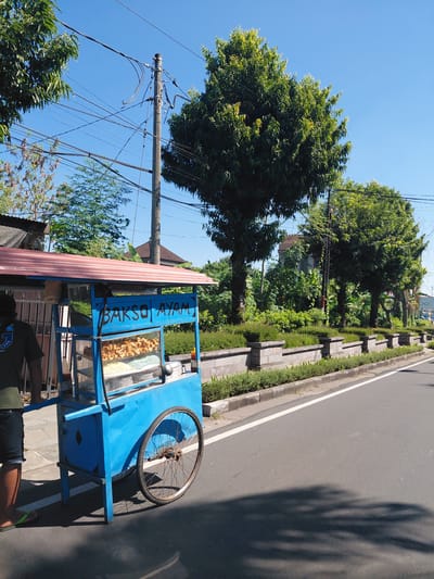This image depicts a sunny roadside scene in Gianyar, Indonesia, featuring a blue and red "Bakso Ayam" (Chicken Meatball) street food cart. A person, whose lower legs and torso are visible, stands behind the cart on the left, wearing a dark shirt, dark shorts, and green flip-flops. The cart has a corrugated red roof and a glass display section showing various items: numerous golden-brown fried snacks on an upper shelf, and multiple metal bowls containing white and light blue noodles on a lower shelf. A small red bottle, likely a condiment, is on the right side of the cart's counter. The cart has a large, spoked wheel with a rusty hub on its right side.
The cart is positioned on a paved asphalt road, which features a solid white line marking its edge. To the right of the road, a concrete curb separates the pavement from a manicured planter area with a low stone wall and green shrubs. Numerous large, green-leafed trees line the road on both sides, with dense foliage obscuring buildings in the background. A tall utility pole with multiple crisscrossing black power lines and cables is prominent against a clear, bright blue sky in the upper-mid section of the image. Strong sunlight casts distinct shadows of the trees and the food cart onto the road surface.