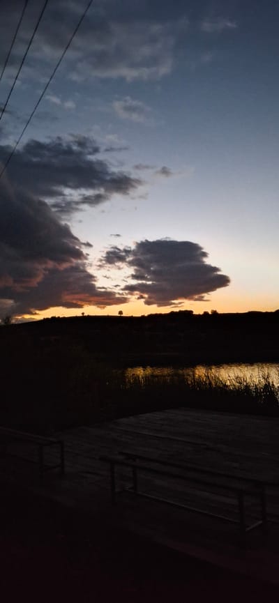 A vertical, low-angle shot captures a dusk scene over the town of Ablanitsa, Bulgaria. In the foreground, a wooden dock or platform with two benches is silhouetted against the fading light. Beyond the dock, tall reeds line the edge of a body of water, reflecting the warm hues of the sunset. The water itself is dark, with shimmering highlights. A dark, silhouetted landscape with rolling hills stretches across the middle ground. A few scattered trees are visible on the horizon, silhouetted against the sky. The sky transitions from a pale blue at the top to warm oranges and yellows near the horizon, with dramatic dark clouds drifting across the upper portion of the frame. Power lines are visible in the upper left corner, crossing diagonally against the sky. The overall atmosphere is serene and tranquil, conveying the end of the day.